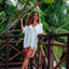 Woman wearing an off-white Ollie Shirt Dress leaning on a wooden railing amid tropical greenery, beach-inspired comfort style.
