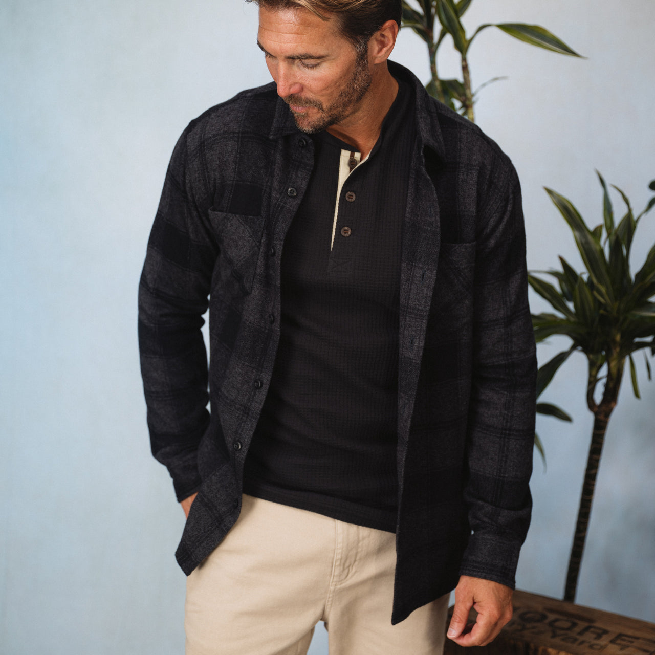 Man wearing a black and gray Reggie Flannel Shirt over a black henley, styled casually with beige pants indoors.