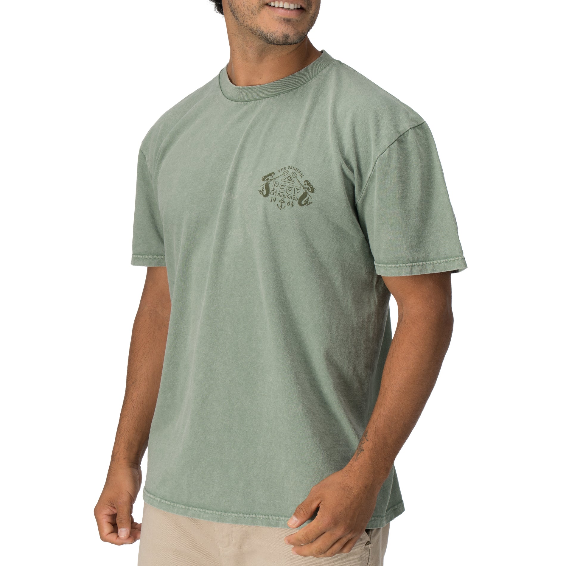 Man wearing a faded green Twining Short Sleeve Tee showing vintage-inspired chest graphic, casual beach apparel.