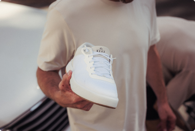 White REEF sneaker held with the toe facing the camera by a seated person