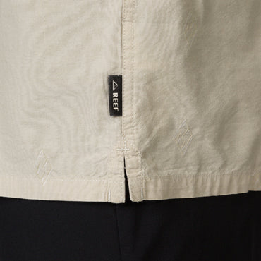 Close-up of the side hem detail on the Harold Camp Shirt showing REEF brand tag on beige, surf-inspired apparel.