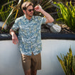 Man wearing Eugene Woven Shirt with blue and white tropical print, standing outdoors by plants and a stone wall adjusting sunglasses