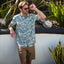 Man wearing Eugene Woven Shirt with blue and white tropical print, standing outdoors by plants and a stone wall adjusting sunglasses