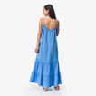 Woman wearing a blue Lainey Maxi Dress shown from back with thin straps, flowing tiers, and casual sandals facing right.