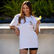 Woman wearing a white Social Boyfriend Crew shirt holding a drink, smiling outdoors with greenery in the background.