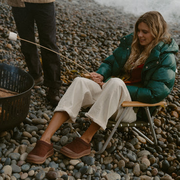 Campfire Lodge brown suede clogs facing left, worn by a woman relaxing beside a beach campfire for cozy comfort