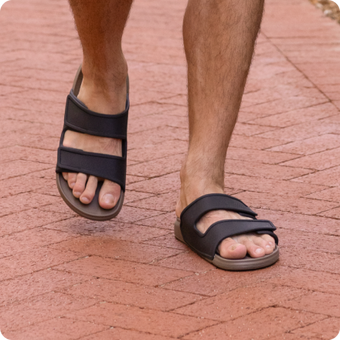 Close up of black two strap sandals facing forward on a person's feet while walking on a brick path