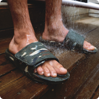 Male feet wearing green camo REEF slide sandals facing forward under outdoor shower water spray on wooden deck