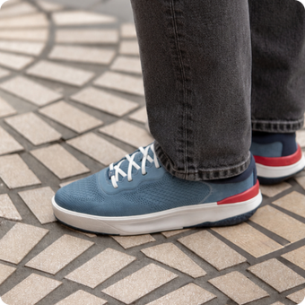 Close-up of right blue sneaker with white laces and red heel detail, worn with dark jeans on tiled floor.