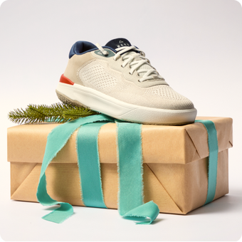 Cream sneaker facing right on a wrapped gift box with teal ribbons and an evergreen branch.
