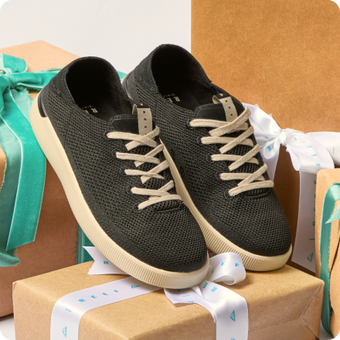 Pair of black knit sneakers with cream laces and soles, toes facing forward, displayed on wrapped gift boxes