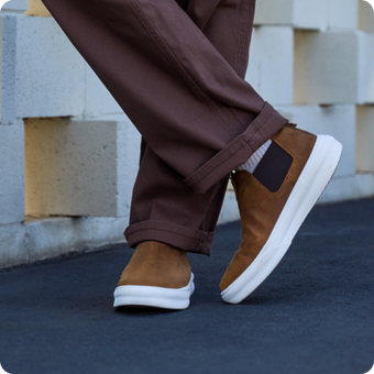 Brown slip-on shoes with white soles facing right, worn with brown pants on pavement