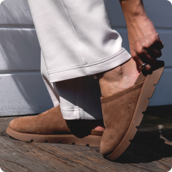 Brown suede slip-on shoes facing left, shown in close-up with textured soles and rolled white pant legs