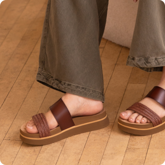Brown platform sandals facing left, with woven toe straps and wide leather bands, worn with wide leg pants on a wood floor