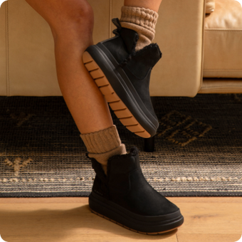 Black ankle boots in side view, toes facing right, with ribbed cuffs and textured rubber soles on a wood floor