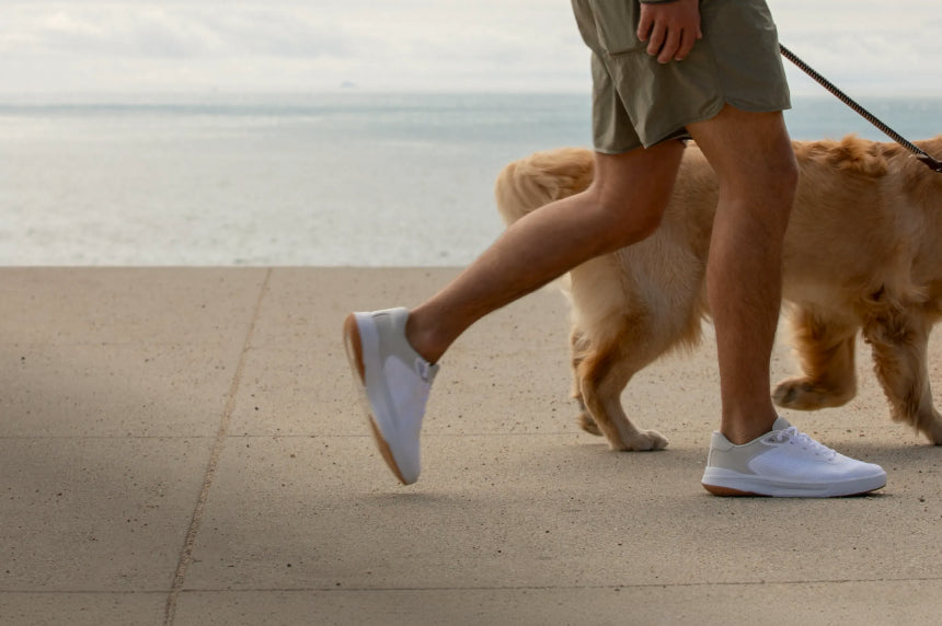 Side view of a person walking a large dog wearing white REEF sneakers with cushioned soles on a beachside path