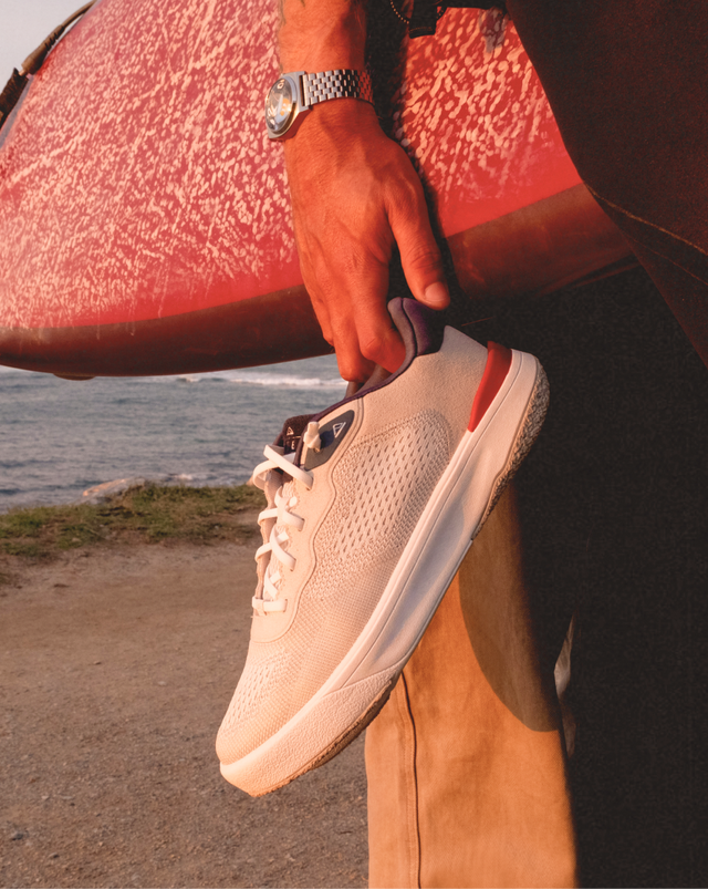 Man holding a White, blue, and red lace-up sneaker by the heel and holding a surfboard with the ocean in the background