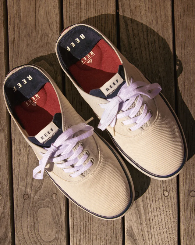 Top-down view of right-facing off-white REEF sneakers with white laces and red cushioned interior on wooden deck