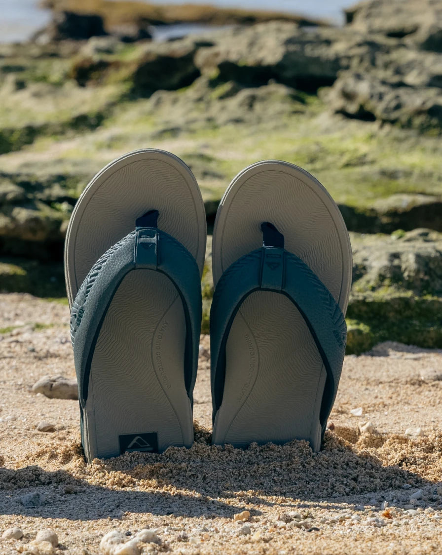 Pair of blue and grey flip flops standing up in the sand at the beach with rocks in the background
