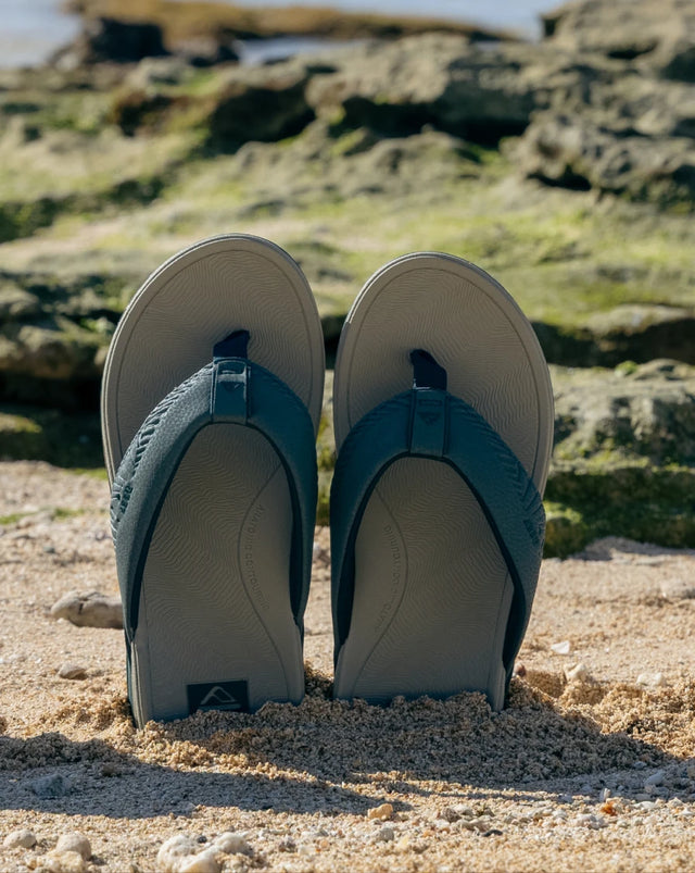 Pair of blue and grey flip flops standing up in the sand at the beach with rocks in the background