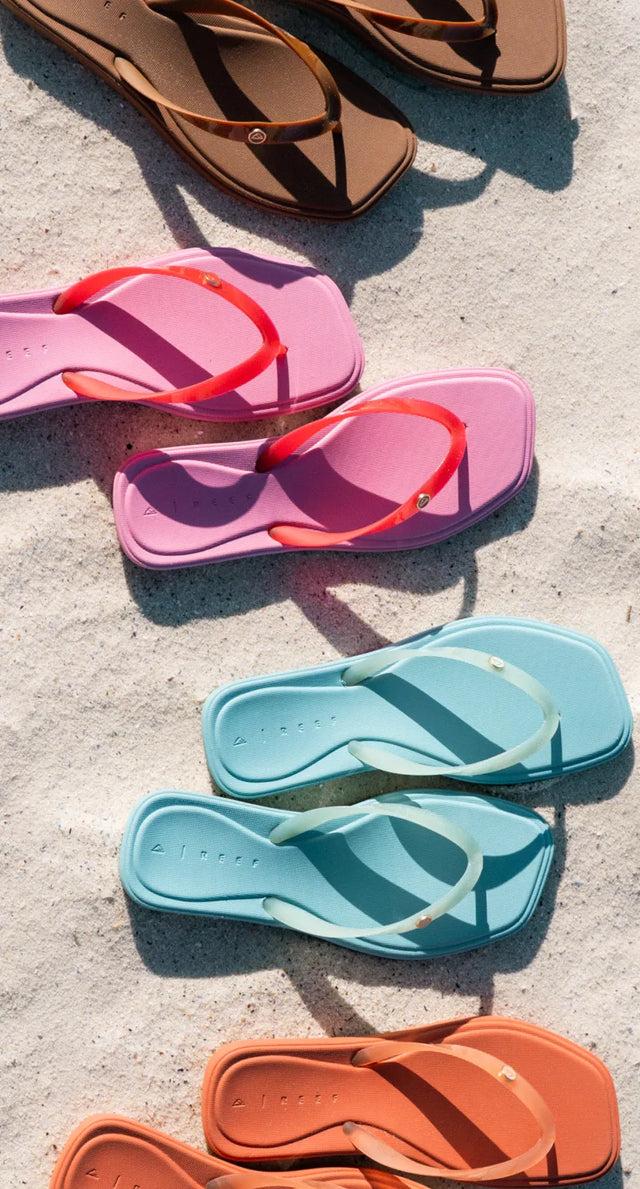 Four pairs of sandals in brown, pink, blue, and orange arranged on textured sand background in bright sunlight.