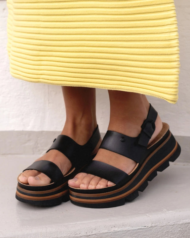 A pair of black and brown platform sandals with a backstrap on a woman's feet, cropped in so the bottom of her yellow dress is showing just above her ankles