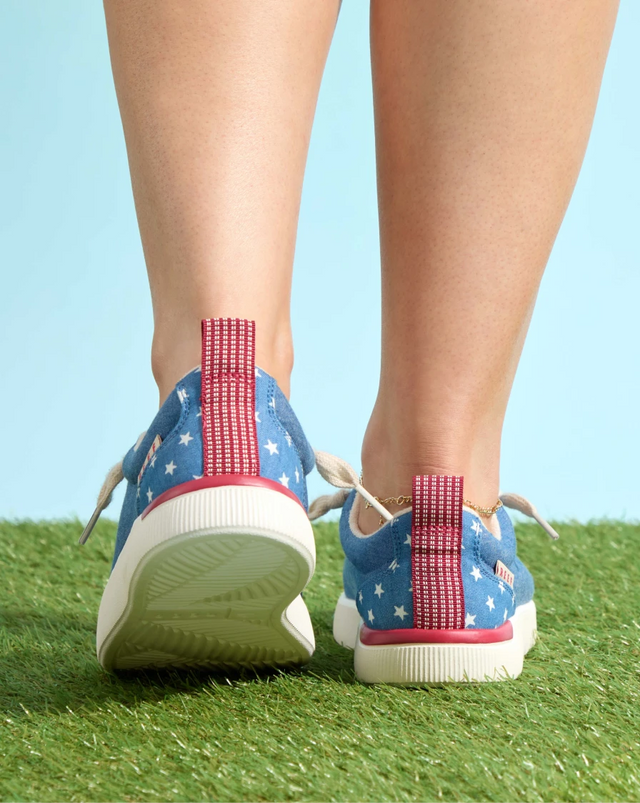 The back of a pair of women's lower legs, wearing some red, white, and blue sneakers with stars on them, and standing on some turf