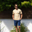 Man wearing a beige Xavier Tee with sunglasses, standing outdoors by plants and wooden fence in bright sunlight