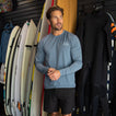 Man wearing Set In Performance Shirt standing indoors near surfboards and wetsuits, showcasing comfort and beach-inspired apparel.