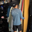 Man wearing a blue Set In Performance Shirt facing away, holding a surfboard in a surf shop with beach gear.