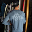 Man wearing a blue Set In Performance Shirt showing back design, standing by surfboards in a beach-inspired setting