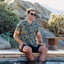Man wearing Kantis Shirt with leaf pattern, seated by pool with legs in water, sunglasses, beach-inspired comfort apparel.