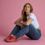 Woman seated against pink backdrop wearing white top, jeans, and red Lay Day Seas shoes facing left, highlighting comfort and style.