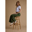 Woman wearing Lay Day Dawn sneakers facing left seated on wooden stool in green pants and white top, beach-inspired comfort shoes.