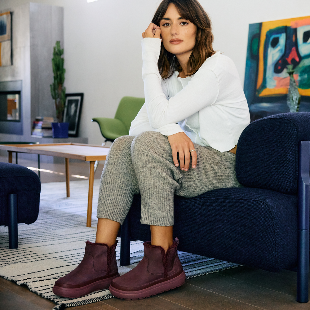 Woman seated on a sofa wearing burgundy ankle boots facing left in a modern living room