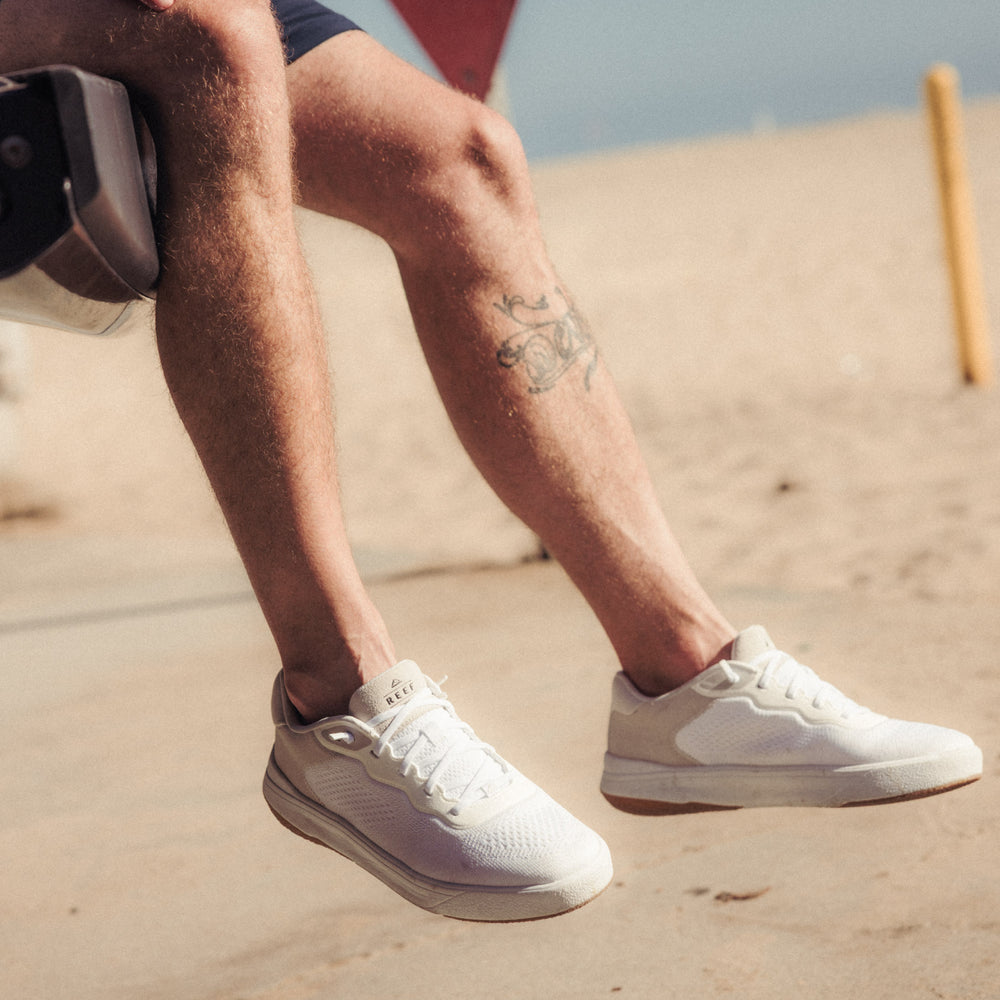 Close-up of lower legs wearing white sneakers facing right on a sandy beach