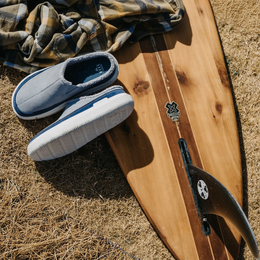 Gray slip-on slippers facing left beside a wooden surfboard and plaid blanket on dry grass
