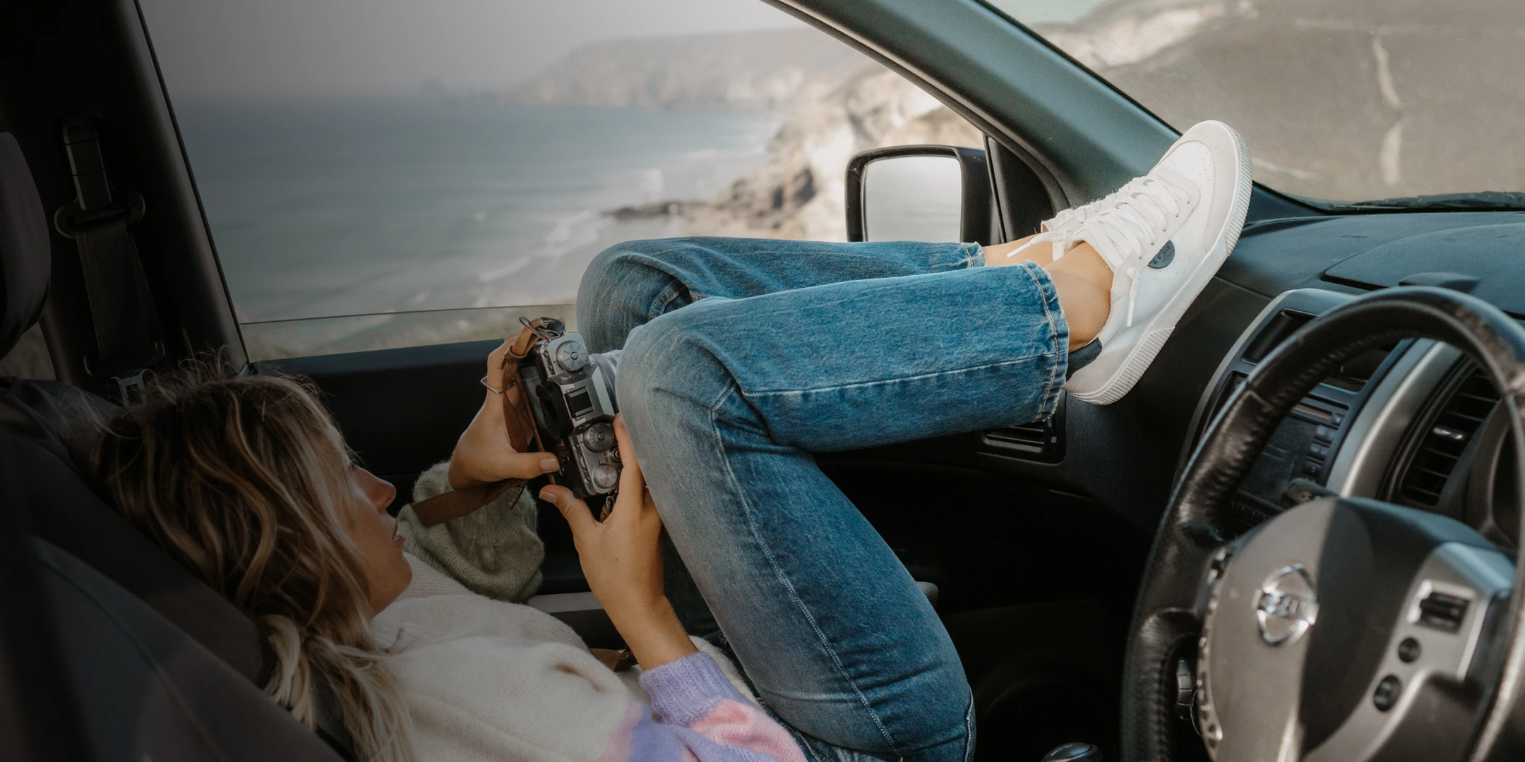 Woman sitting in the driver's seat of a call with a camera, feet up wearing the Lay Day Sneaker.