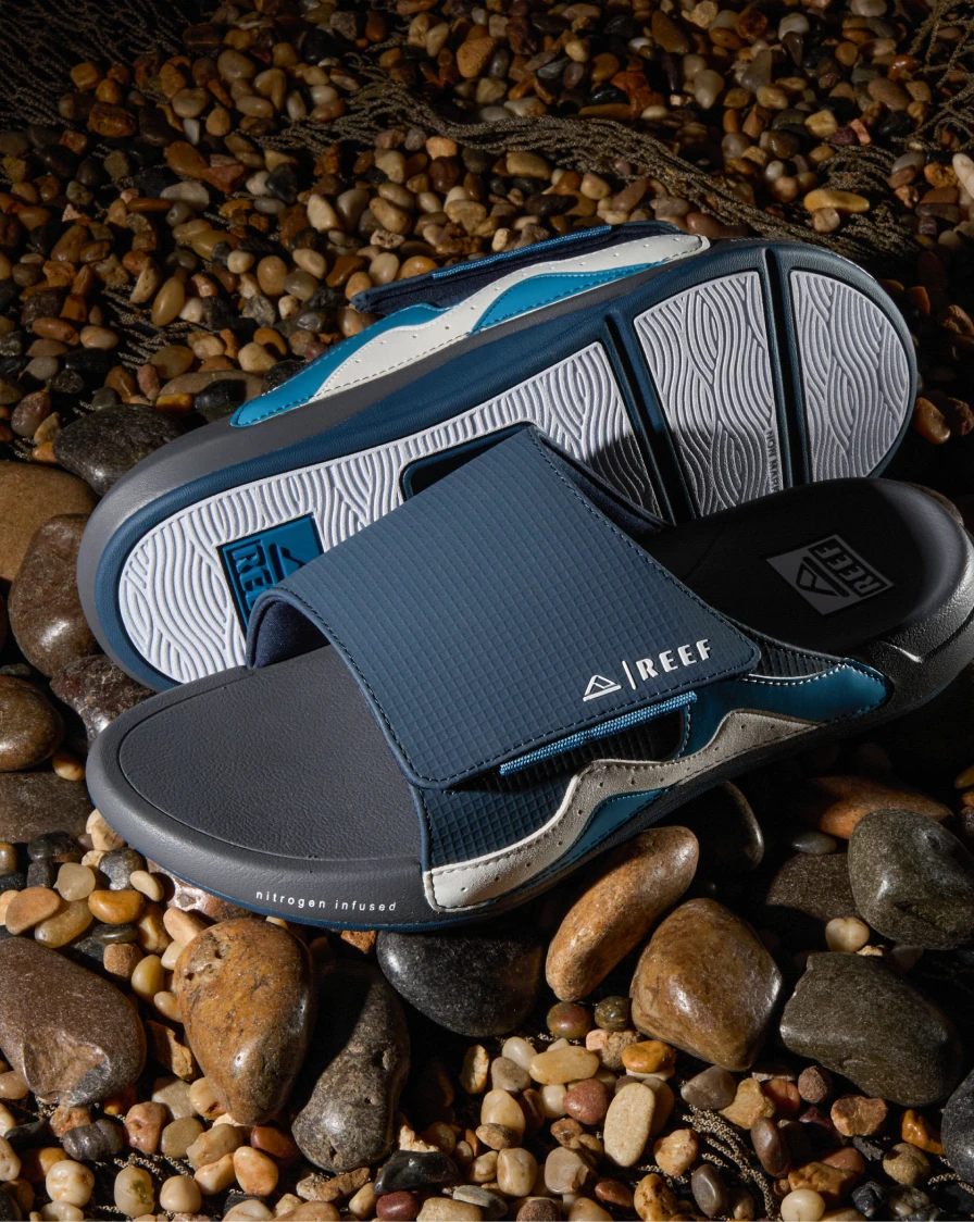 Pair of blue, white, and grey single-strap slide sandals sitting on some small pebbles