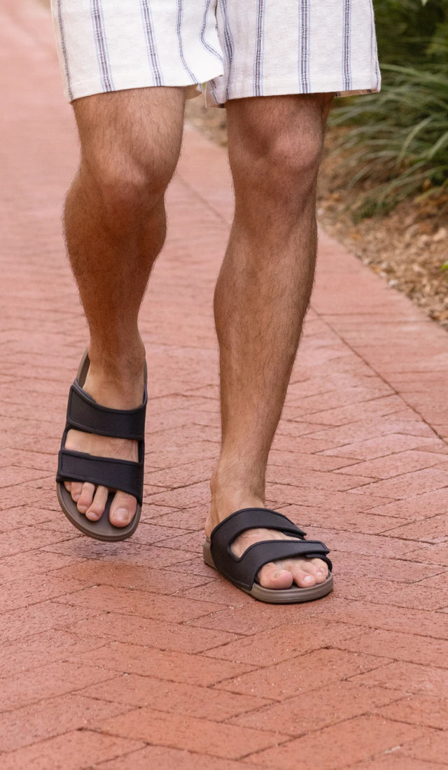 Man's legs walking on a brick path wearing some brown two-bar slide sandals and some white shorts with plants in the background