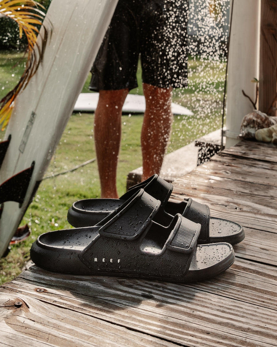 Pair of black two-bar slide sandals on a wooden deck with a person holding a surfboard under an outdoor shower in the background.