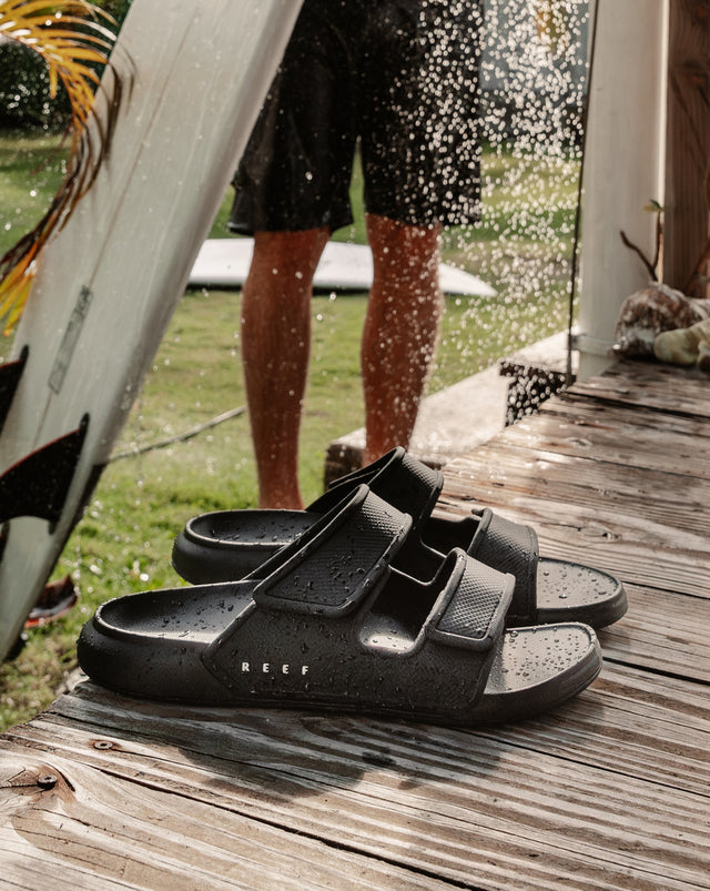 Pair of black two-bar slide sandals on a wooden deck with a person holding a surfboard under an outdoor shower in the background.