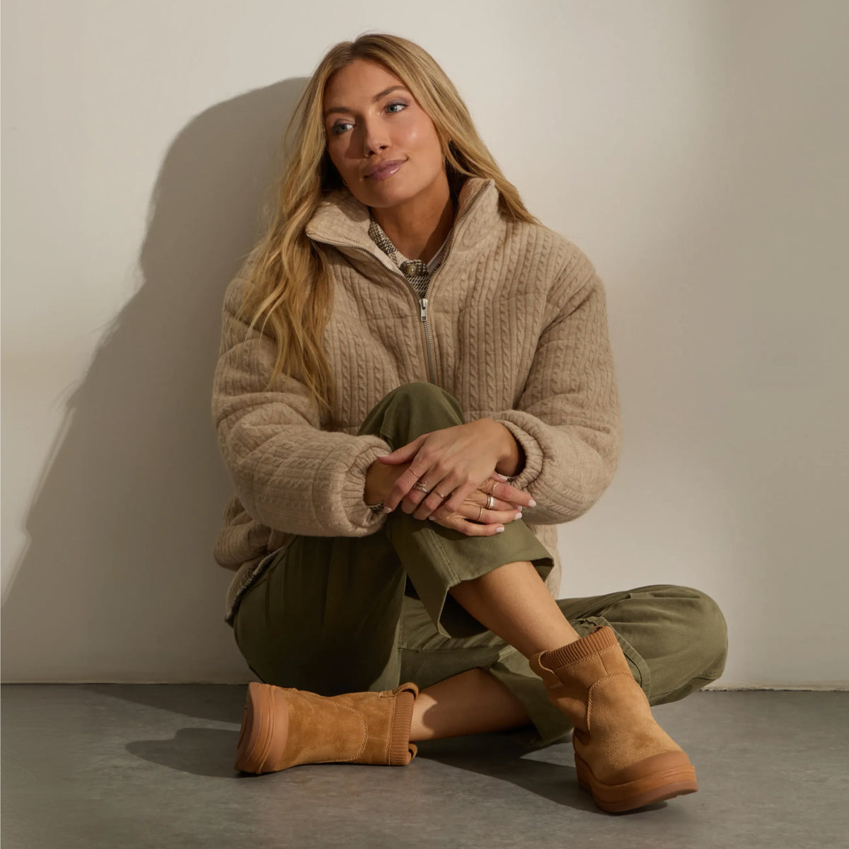 Woman seated against a wall wearing tan ankle boots with toes facing forward and slightly outward