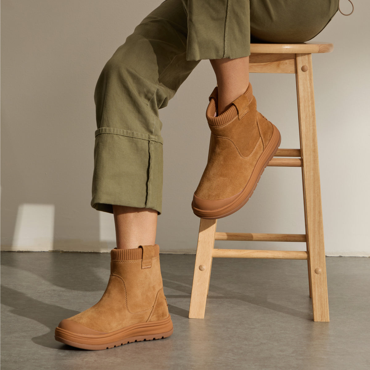 Person wearing tan ankle boots, both facing left, with one boot lifted beside a wooden stool.