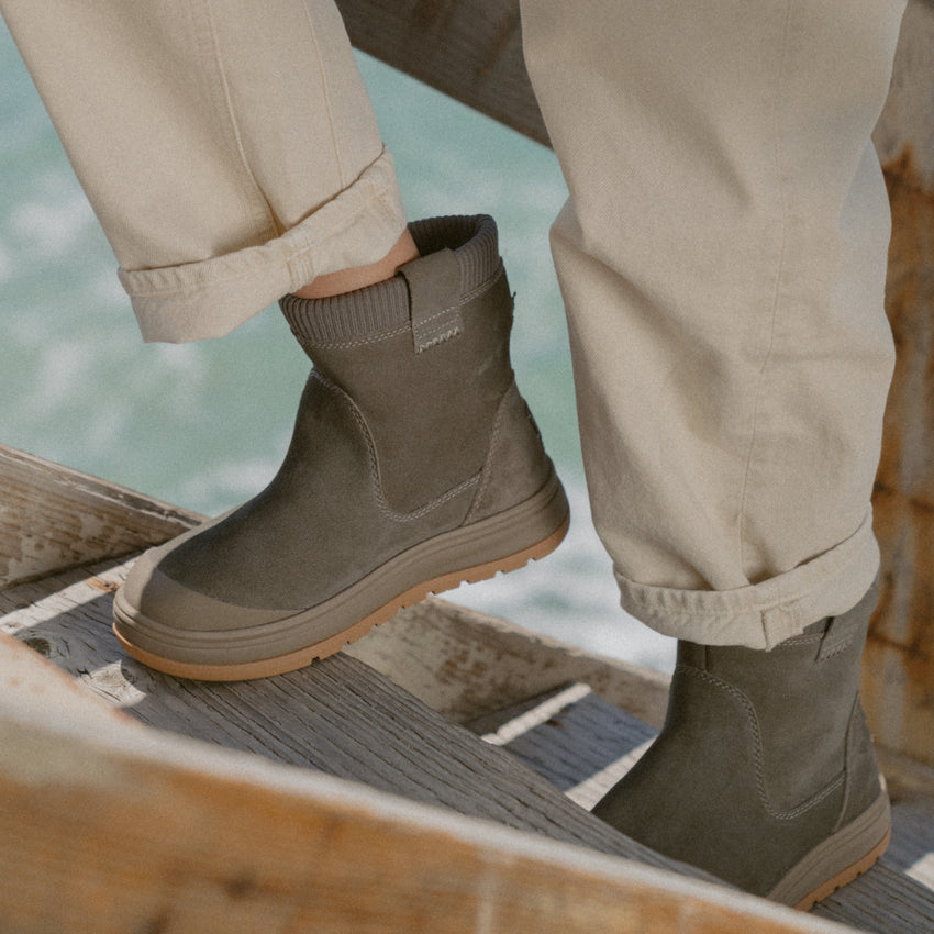 Close-up of brown ankle boots, one facing left and one facing right, worn on a wooden boardwalk over water