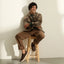 Smiling man seated on a wooden stool, wearing brown lace-up shoes facing left