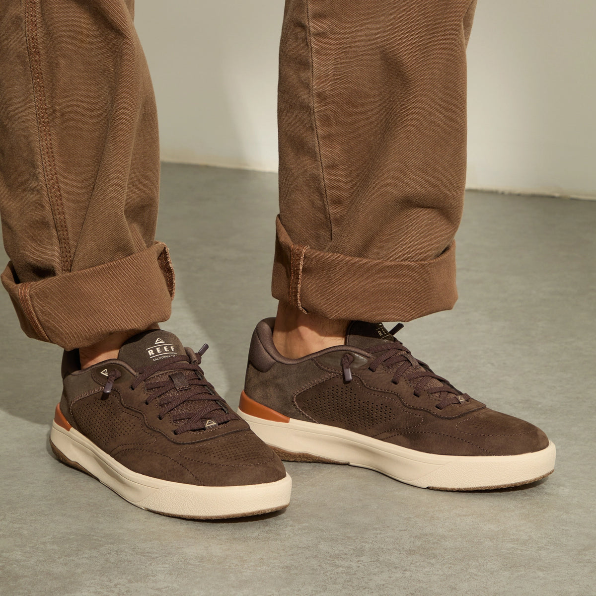Pair of brown sneakers facing right, worn with cuffed brown pants on a concrete floor