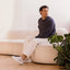 Man seated on a sofa wearing white sneakers with toes facing left, in a bright room with a leafy plant