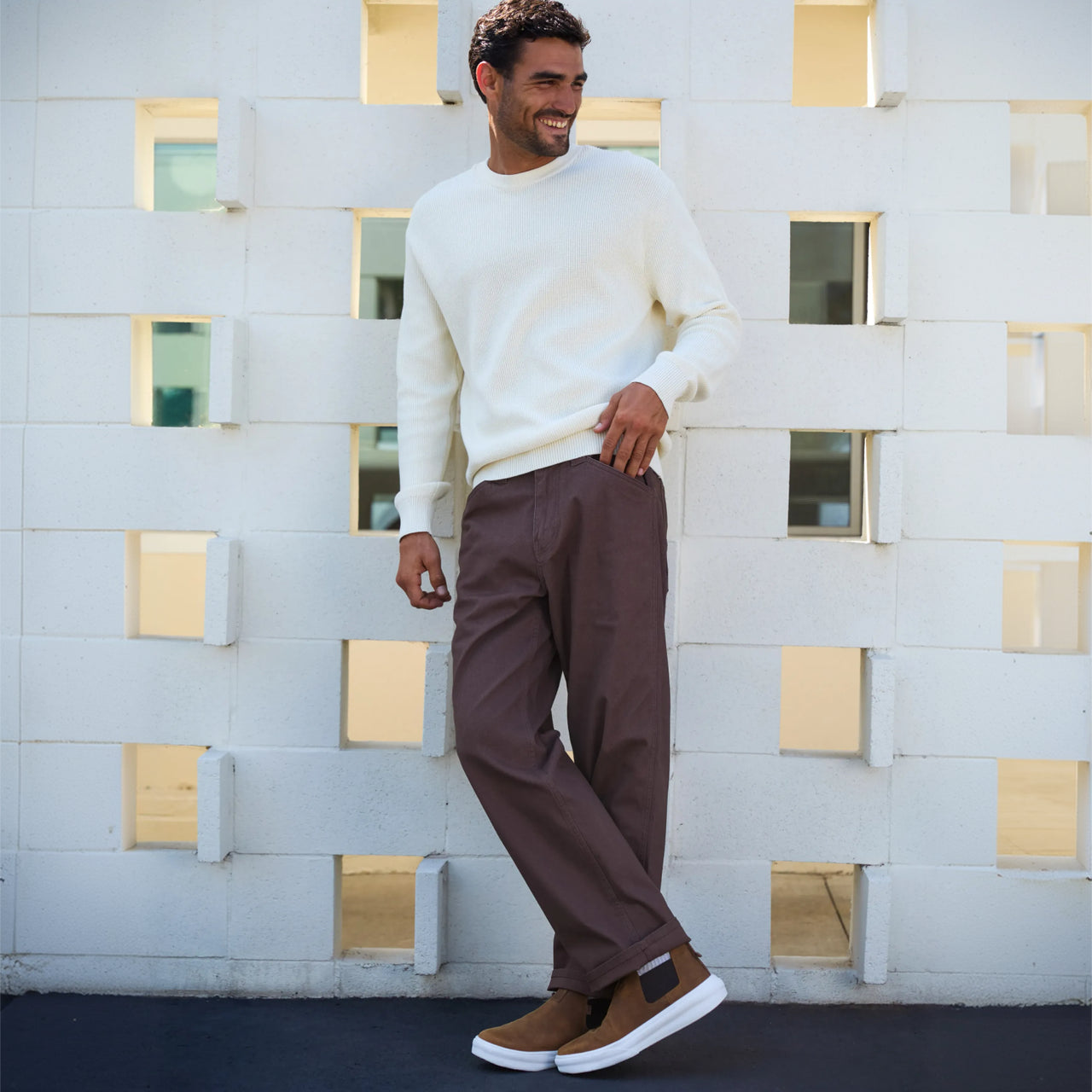 Man wearing brown Evo footwear facing right, styled with white sweater and brown pants against white wall background.
