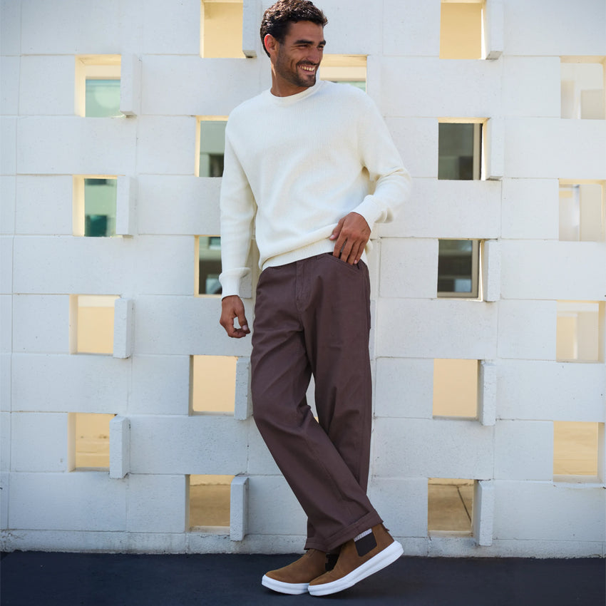Man wearing brown Evo footwear facing right, styled with white sweater and brown pants against white wall background.